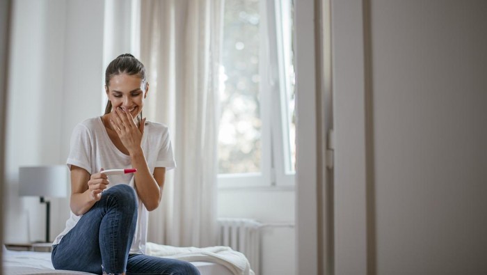 Image of a smiling Asian woman holding a pregnancy test kit and looking at the test result