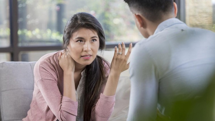 While in a disagreement, a young couple discusses problems. The wife sits on the sofa and gestures in frustration while husband listens intently. The discussion is taking place in a living room.