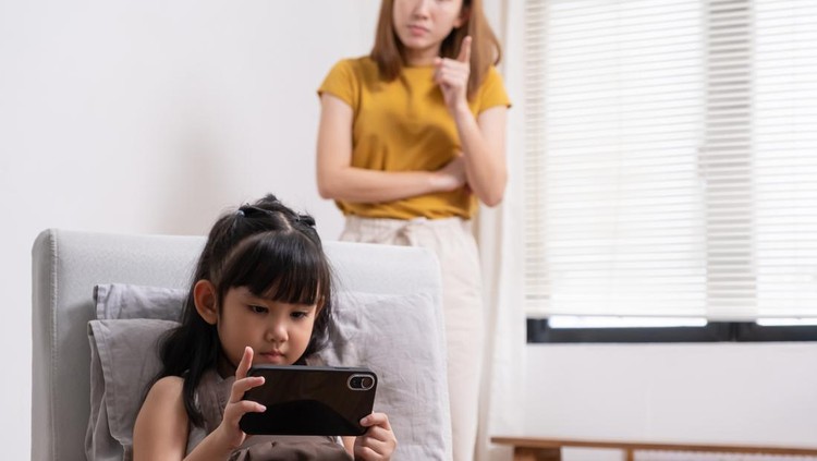 Young Asian girl looking at her smartphone while watching video or playing game online with mother warning her to stop at the back ground. Social network or smartphone addict in kids and children.