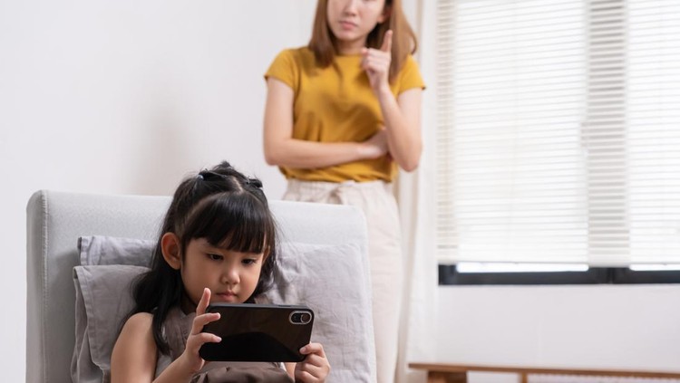 1429402981 Young Asian girl looking at her smartphone while watching video or playing game online with mother warning her to stop at the back ground. Social network or smartphone addict in kids and children.