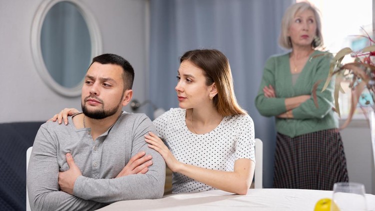 Loving young woman supporting and comforting her upset stressed husband while her mother-in-law arguing on background