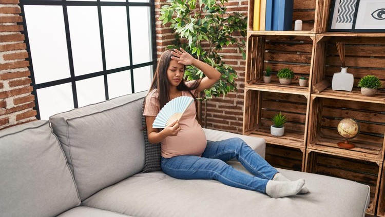Young latin woman pregnant using hand fan sitting on sofa at home