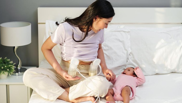 happy mother using breast pump machine to pumping milk while talking with her newborn baby on a bed