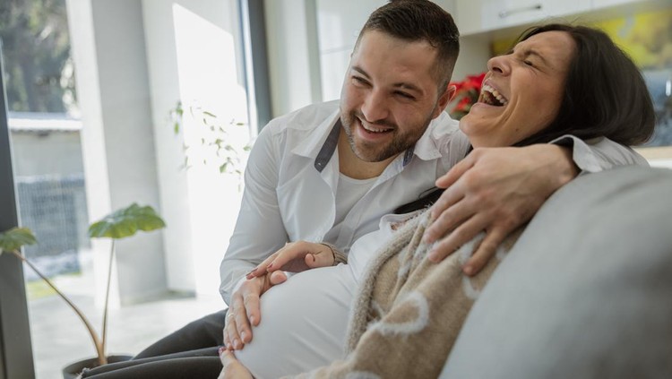 Man touching belly of laughing pregnant woman sitting on sofa at home