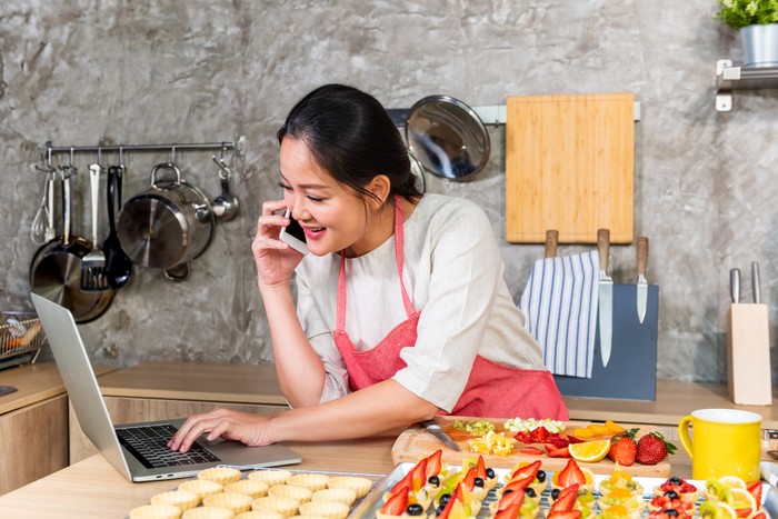 Asian woman making fruit tart for online selling delivery. Female small business bakery shop owner entrepreneur working in laptop computer and mobile phone get order for customer. Small business work at home concept.