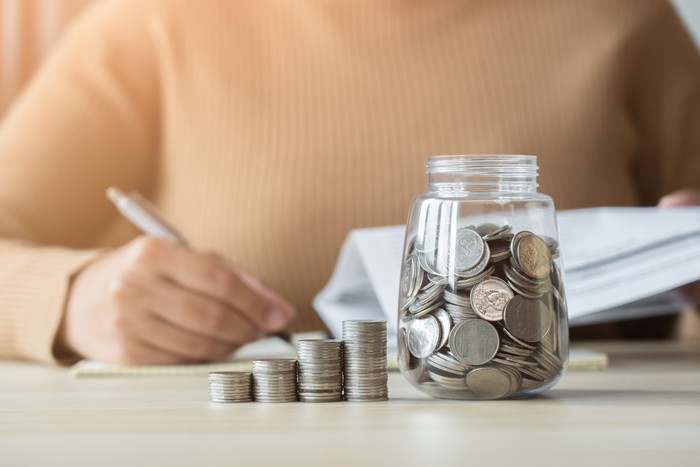 Woman with coins jar taking note to checklist expenses. Concept of doing accounting plan with coin jar and woman writing in a book.