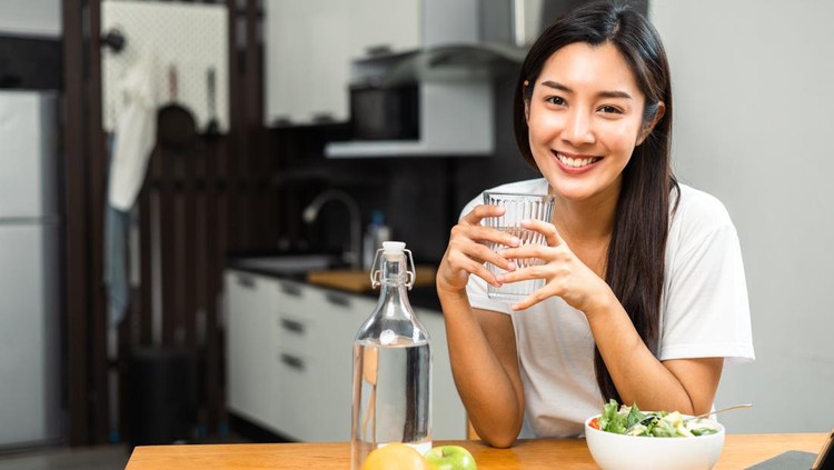 Young female drinking water in glass for refreshing for diet and wellness on table. Beautiful woman happily drink fresh water before breakfast in kitchen in the morning. Healthy food concept.