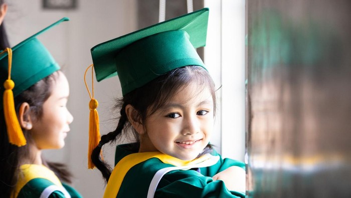 Asian elementary age girls wearing graduation caps and gowns smile while standing inside school.