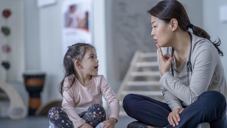 A female therapist sits beside her young patient on the floor as they work on some speech therapy exercises.  They are each dressed casually and the therapist has her hand on her chin as she works to help the young girl through phonetical sounds.
