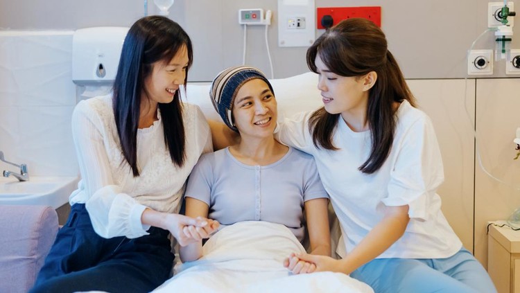 Happy female patient visited by friends in hospital ward, sitting beside on the bed and giving support for recovery
