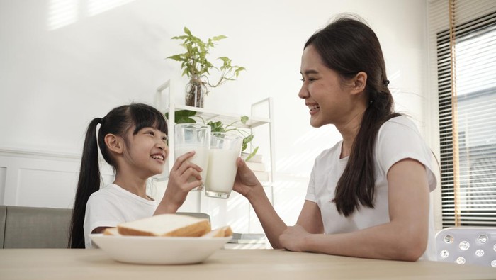 A Healthy Asian Thai family, little daughter, and young mother drink fresh white milk in glass and bread joy together at a dining table in morning, wellness nutrition home breakfast meal lifestyle.