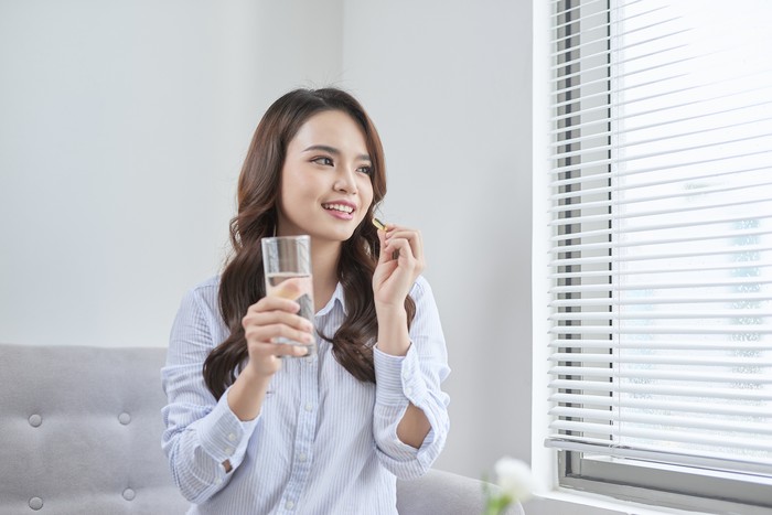 Sick Asian woman eating pills with a glass of water in hand near window in her house.
