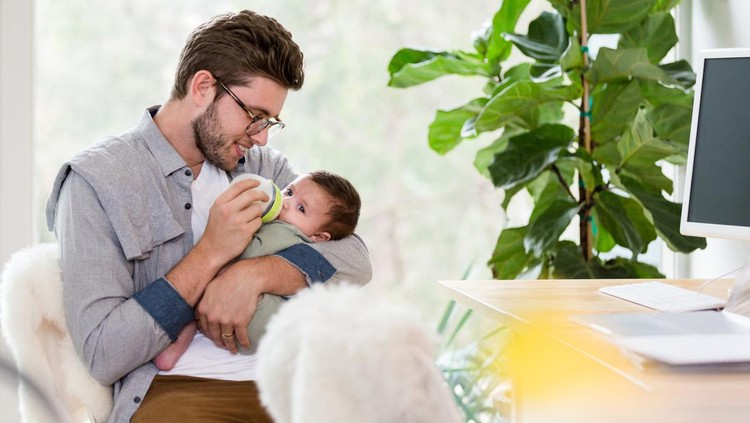 As he works from his home, a young dad gives his newborn son a bottle. The man is sitting at a desk in his home office.