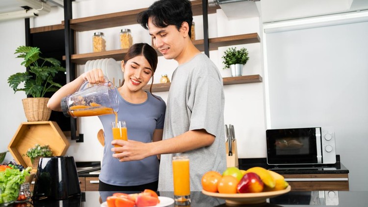 Asian woman pouring smoothies for a man. Happy healthy Asian couple using a blender machine for making healthy vegan smoothies. Couple making vegan smoothies together at home for a healthy lifestyle.