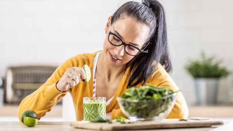 A smiling brunette is preparing a spinach smoothie and flavoring it with lime juice.
