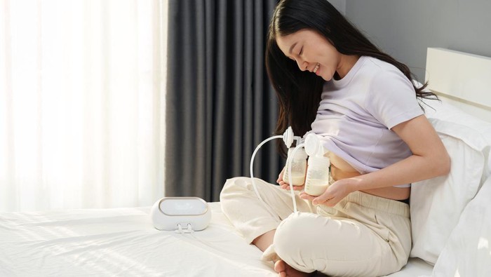 mother using breast pump machine to pumping milk with infant baby on a sofa in the living room at night