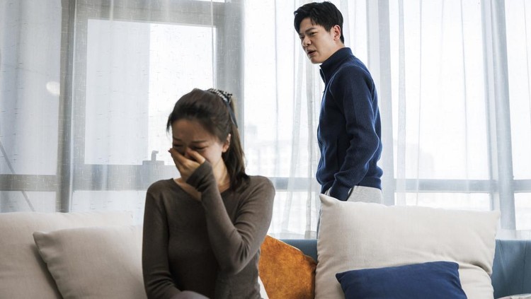 This is a young asian couple. They wear casual clothes. Sitting on the sofa in the living room at home. There was a quarrel between them, and the man was very excited. Women are sad.