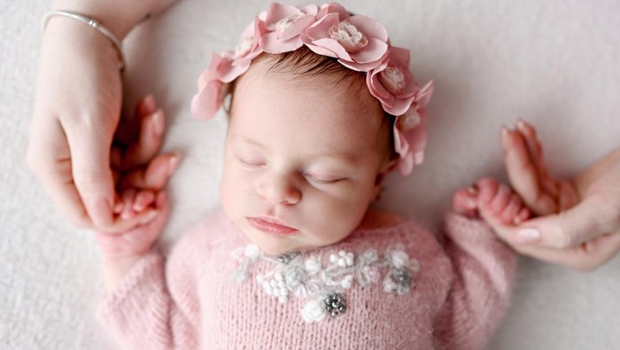 Newborn baby girl wearing flower wreath sleeping and mother holding her hands. Infant child kid napping, parent care and love