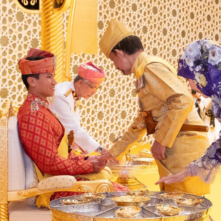 <p>Di upacara kali ini, anggota keluarga akan mengoleskan benda seperti pasta bedak yang disebut Bedak Diraja ke tangan mempelai pria dan wanita. Dalam potret yang dibagikan, Sultan Haji Hassanal Bolkiah tampak melakukan prosesi tersebut didampingi oleh sang istri, Saleha binti Mohamed Alam. (Foto: Instagram @pelitabrunei)</p>