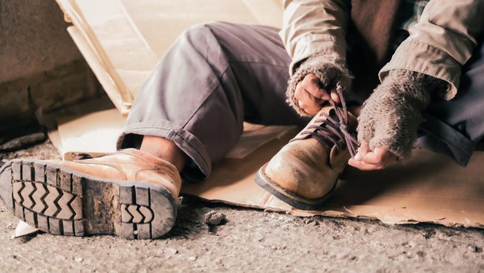 Senior man homeless wearing sweater and cloth gloves with sitting on cardboard and tying laces on shoes. Close up of asian man homeless in shelter and wear leather shoes boot and tie lace his shoes.