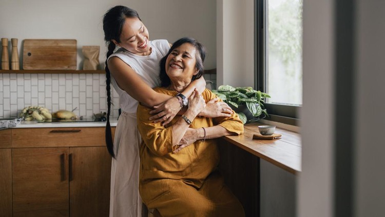 A smiling Asian woman enjoying drinking tea while her loving daughter is embracing her.