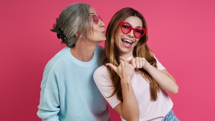 Playful mother and adult daughter in funky eyeglasses having fun against pink background