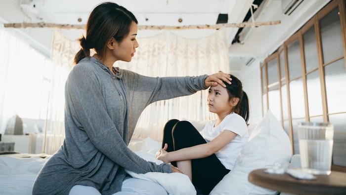 Sick little daughter lying in bed. A loving Asian mother checking temperature on her forehead. A glass of water and medicine on the side table