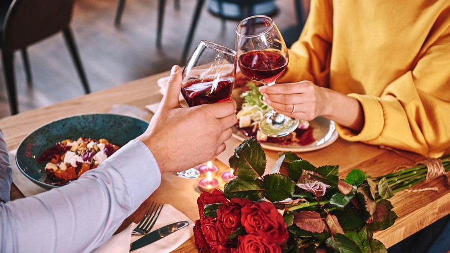 Happy moments. Close-up of couple holding red wine in glasses in restaurant. Red roses and candles are lying on the table near the red wine. Young woman in mustard sweater looks at her boyfriend