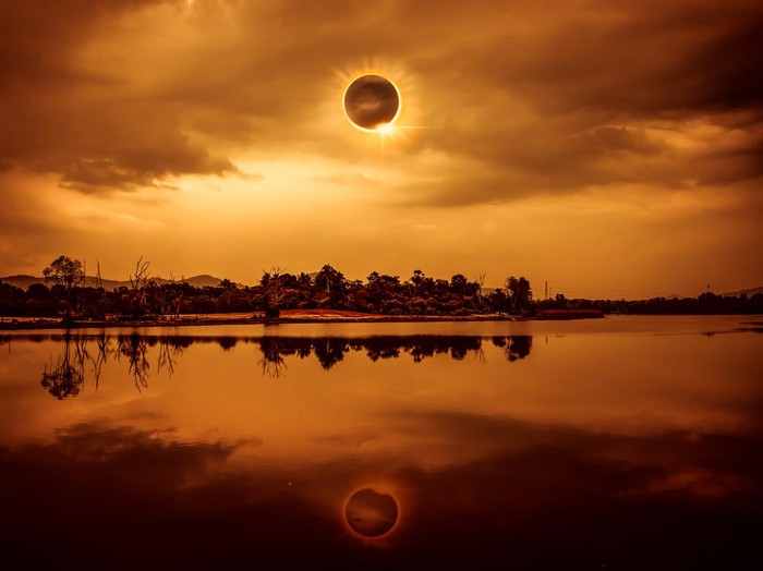 Amazing scientific natural phenomenon. The Moon covering the Sun. Total solar eclipse with diamond ring effect glowing on sky above silhouettes of trees, river area. Serenity nature background.