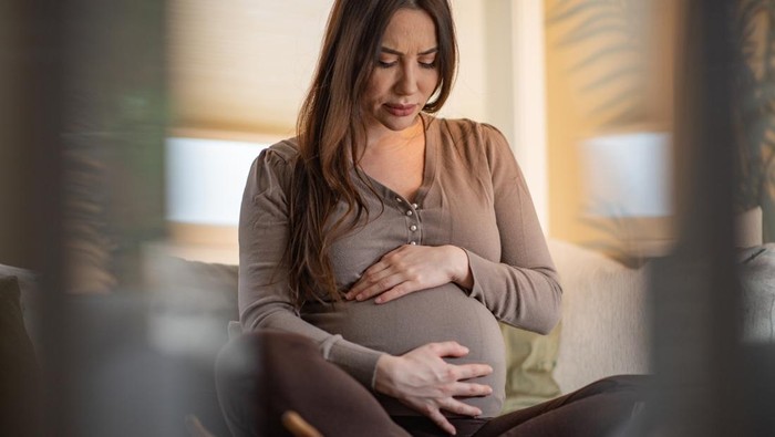 Pregnant young woman grimacing with hands on stomach while sitting at home