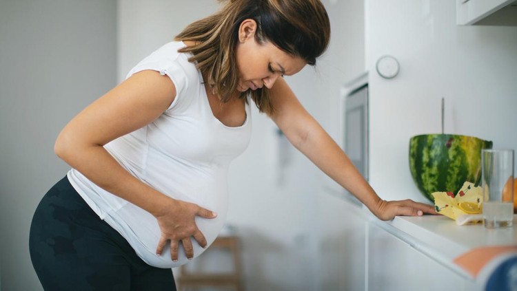 Pregnant young woman grimacing with hands on stomach while sitting at home