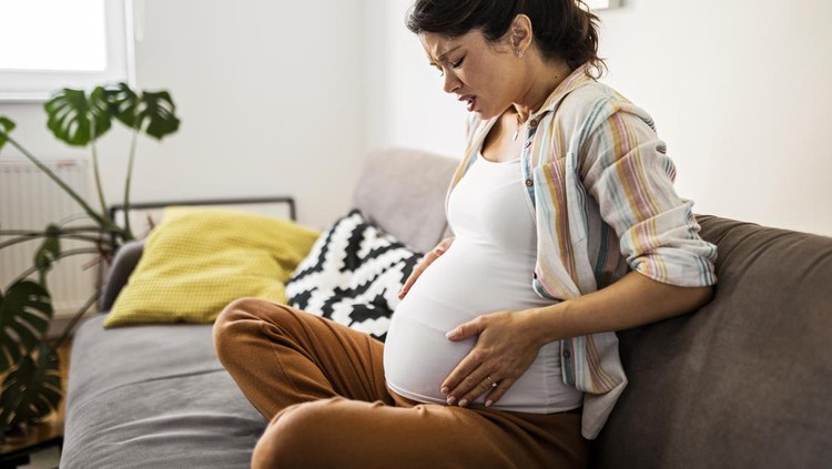 Pregnant young woman grimacing with hands on stomach while sitting at home