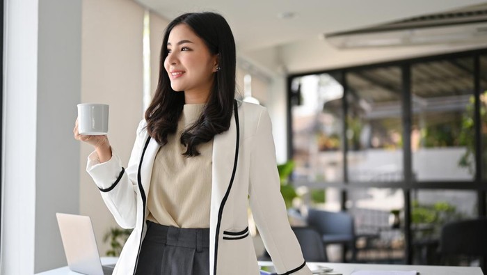 Attractive and successful millennial Asian businesswoman in white suit stands in her office looking out the window and daydreaming about her career success while sipping coffee.