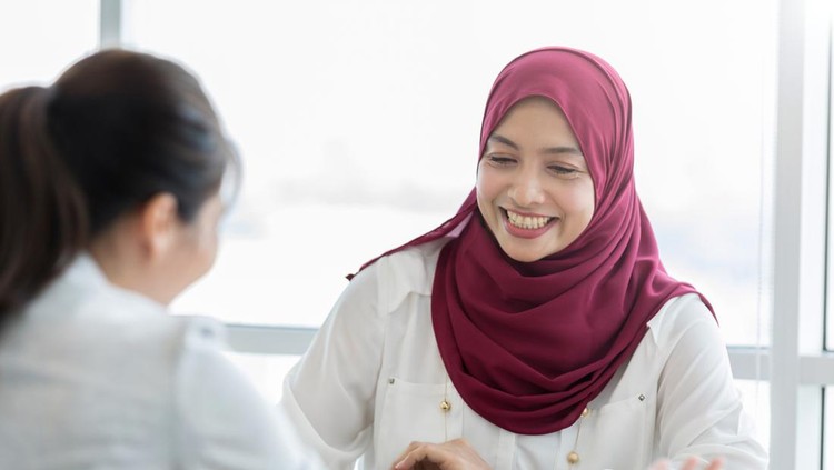 A smiling young Malaysian businesswoman sits across her office desk from an unrecognizable coworker.  She looks down as they discuss a project.