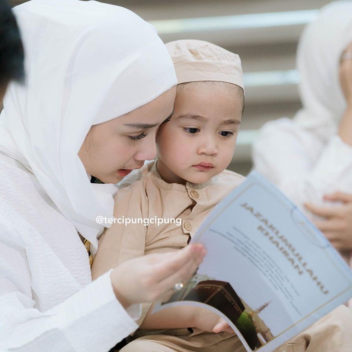 <p>Rayyanza terlihat tampan mengenakan baju koko warna krem. Ia terduduk tenang di atas pangkuan sang Bunda sambil melihat buku panduan tentang ibadah haji. (Foto: Instagram: @tercipungcipung)</p>