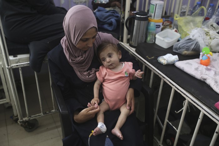 Abdulrahman Al-Rai holds his daughter, Hana Al-Rai, 3, who suffers from diabetes, a weakened immune system, and malnutrition,  as she gets help from her sister to test her blood sugar at Al-Aqsa Martyrs Hospital in Deir al-Balah in the central Gaza Strip, where she is receiving treatment, Saturday, June 1, 2024. The family is displaced from the northern Gaza Strip. (AP Photo/Jehad Alshrafi)