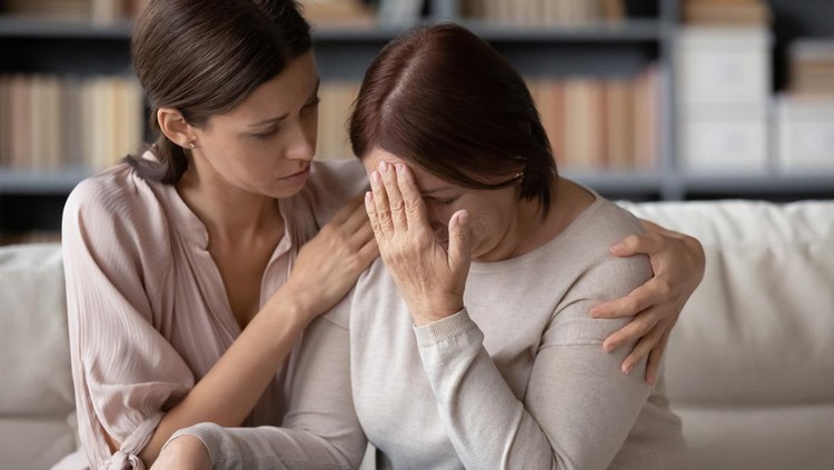 Close up caring grownup daughter comforting unhappy mature mother, embracing, helping to overcome problems, sitting on couch at home, unhealthy elderly mum touching forehead, suffering from headache