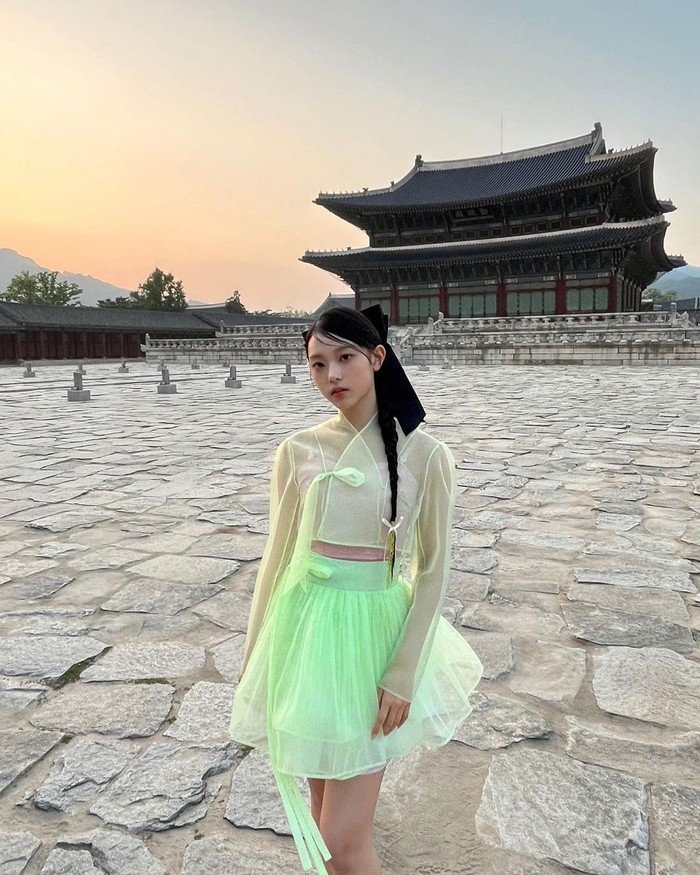 Haerin tampil feminin dan cute dalam outfit hanbok modern berwarna hijau neon transparan yang menyala. Ia memusatkan tampilannya dengan gaya rambut kepang dan pita hitam yang khas. / Foto: Instagram/newjeans_official