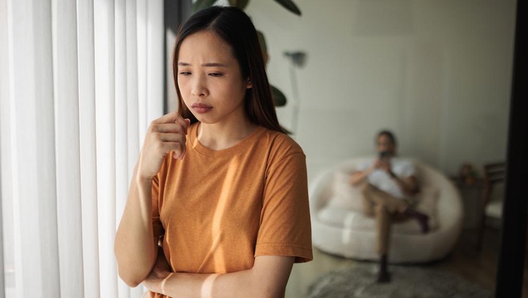 A young woman looks worried while her husband sits on the sofa in the background.