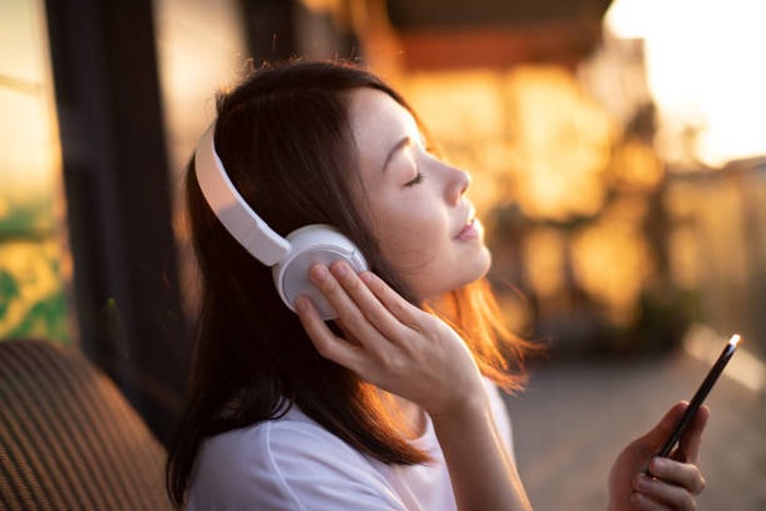 Portrait of smiling young Asian woman with eyes closed enjoying music over headphones and using smartphone, relaxing on the balcony against sunlight.