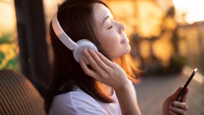 Portrait of smiling young Asian woman with eyes closed enjoying music over headphones and using smartphone, relaxing on the balcony against sunlight.