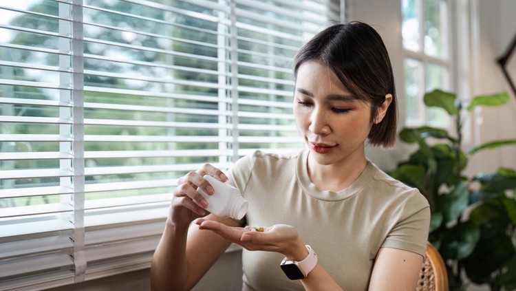 wanita minum obat High angle view, Asian teenage woman pours medicine into her hand while sitting on a sofa in the living room. Healthcare and medicine concept