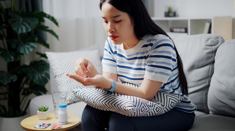 High angle view, Asian teenage woman pours medicine into her hand while sitting on a sofa in the living room. Healthcare and medicine concept