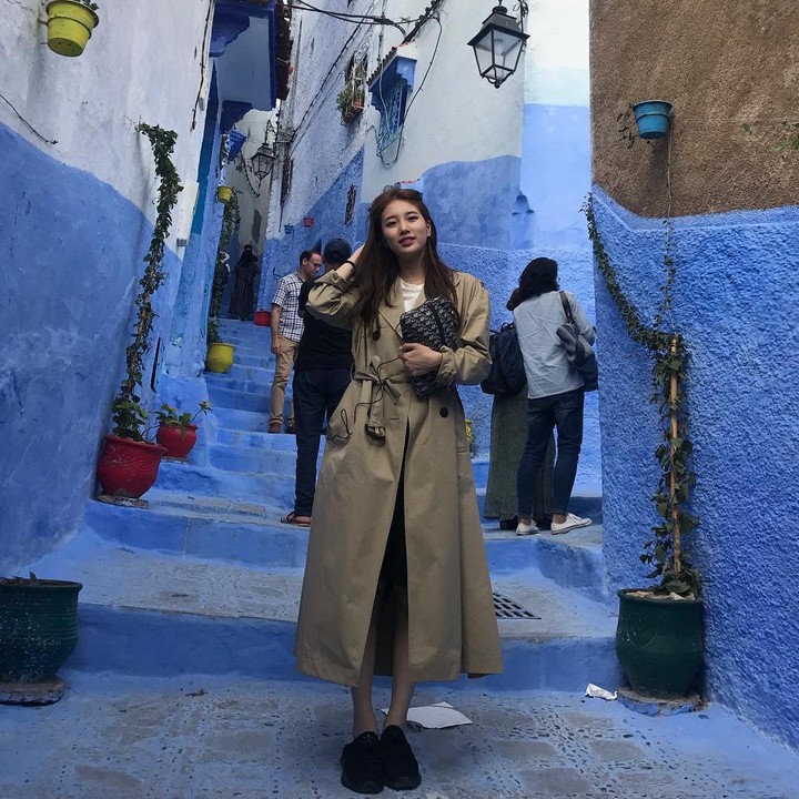 <p>Ketika berkunjung ke Maroko, Bae Suzy tampil modis dengan <em>trench coat</em> warna cokelat yang kontras dengan latar belakang Chefchaoue yang serba biru. (Foto: Instagram @skuukzky)</p>