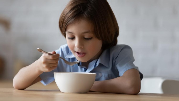 Little hungry cute boy sit at dinner table in kitchen holds spoon eating porridge or whole grain dry cereal with pleasure and appetite, enjoy balanced food. Nutrition, tasty healthy breakfast concept