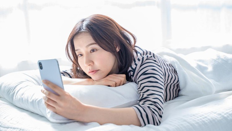 A woman falling asleep staring at a smartphone screen in bed.
