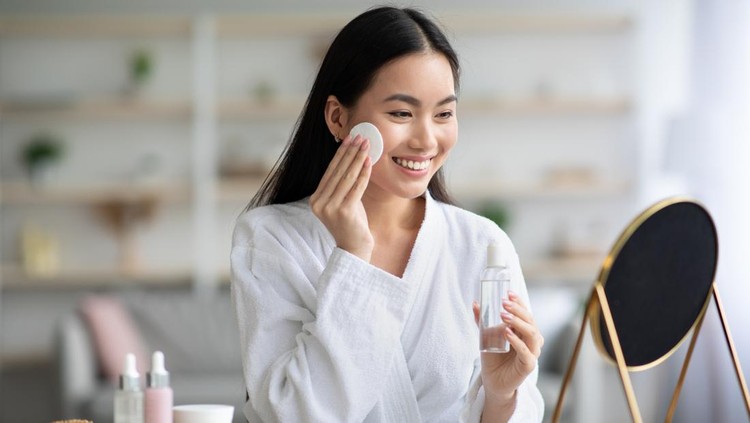 Cheerful asian woman cleaning her face&comma; using cotton pads and cleansing product&comma; looking at mirror in bedroom&period; Young attractive korean lady using face toner and cotton pad&comma; home interior&comma; empty space
