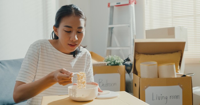 Young woman moving house sit on couch eating instant noodles after unpacking.