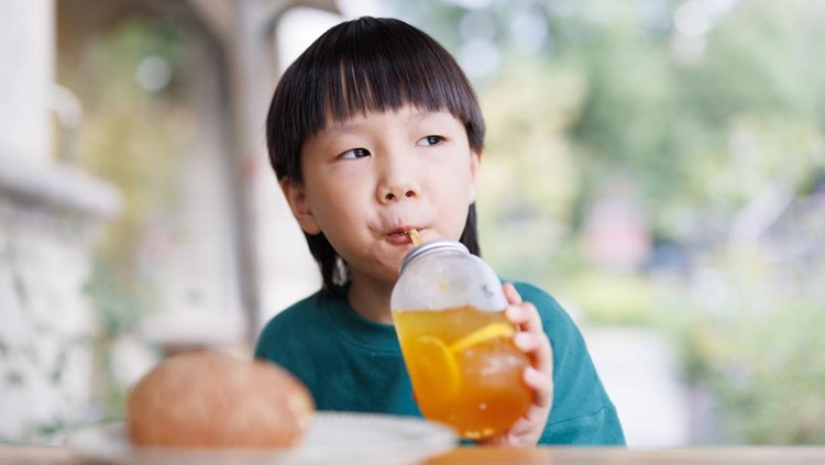 An Asian boy, about 5 years old. Black haired, dressed in casual clothing, sitting outdoors, eating bread and drinking juice.