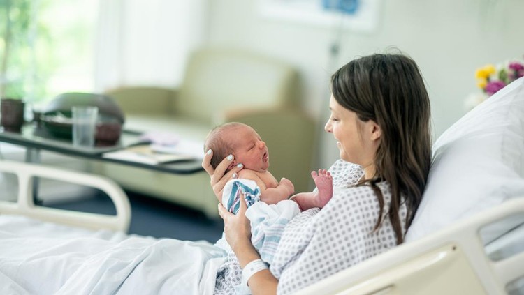 A new Mother sits up in her hospital bed shortly after delivery as she holds her newborn out in front of her and studies his features.  She is wearing a hospital gown and is laying in her hospital bed with the inafnt.